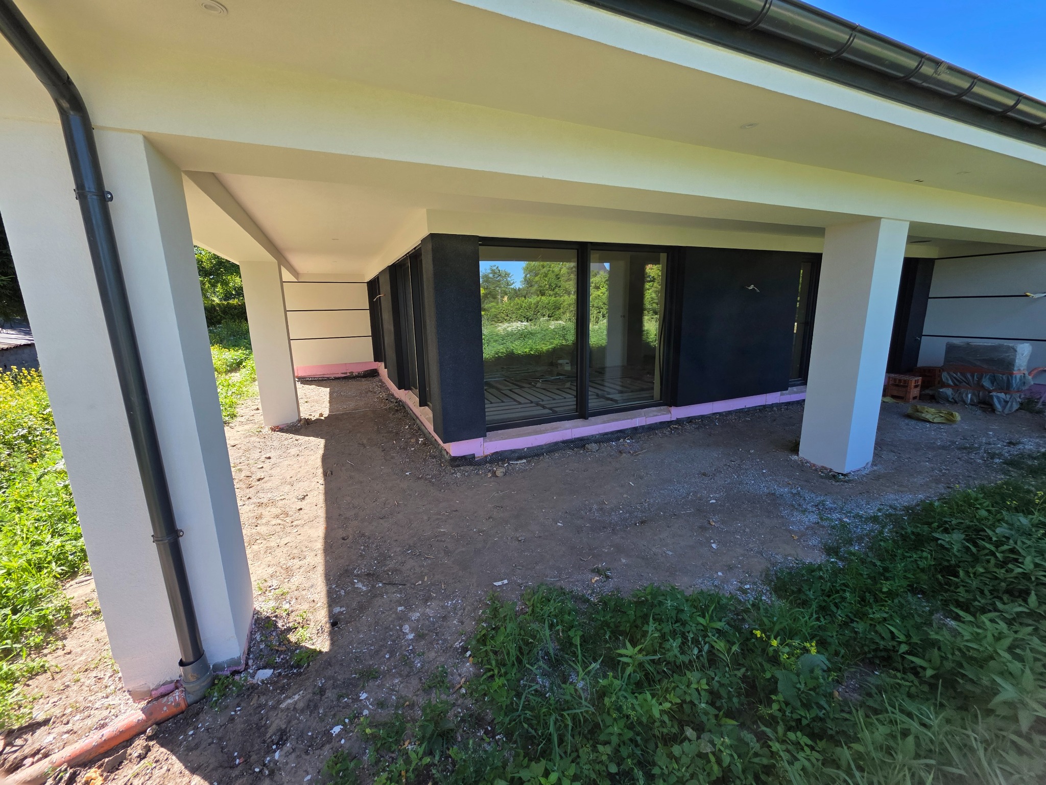 Częściowo ocieplony budynek z widoczną warstwą izolacji (różowy styropian) przy podstawie ścian i dużymi oknami. Widoczne kolumny podtrzymujące dach.