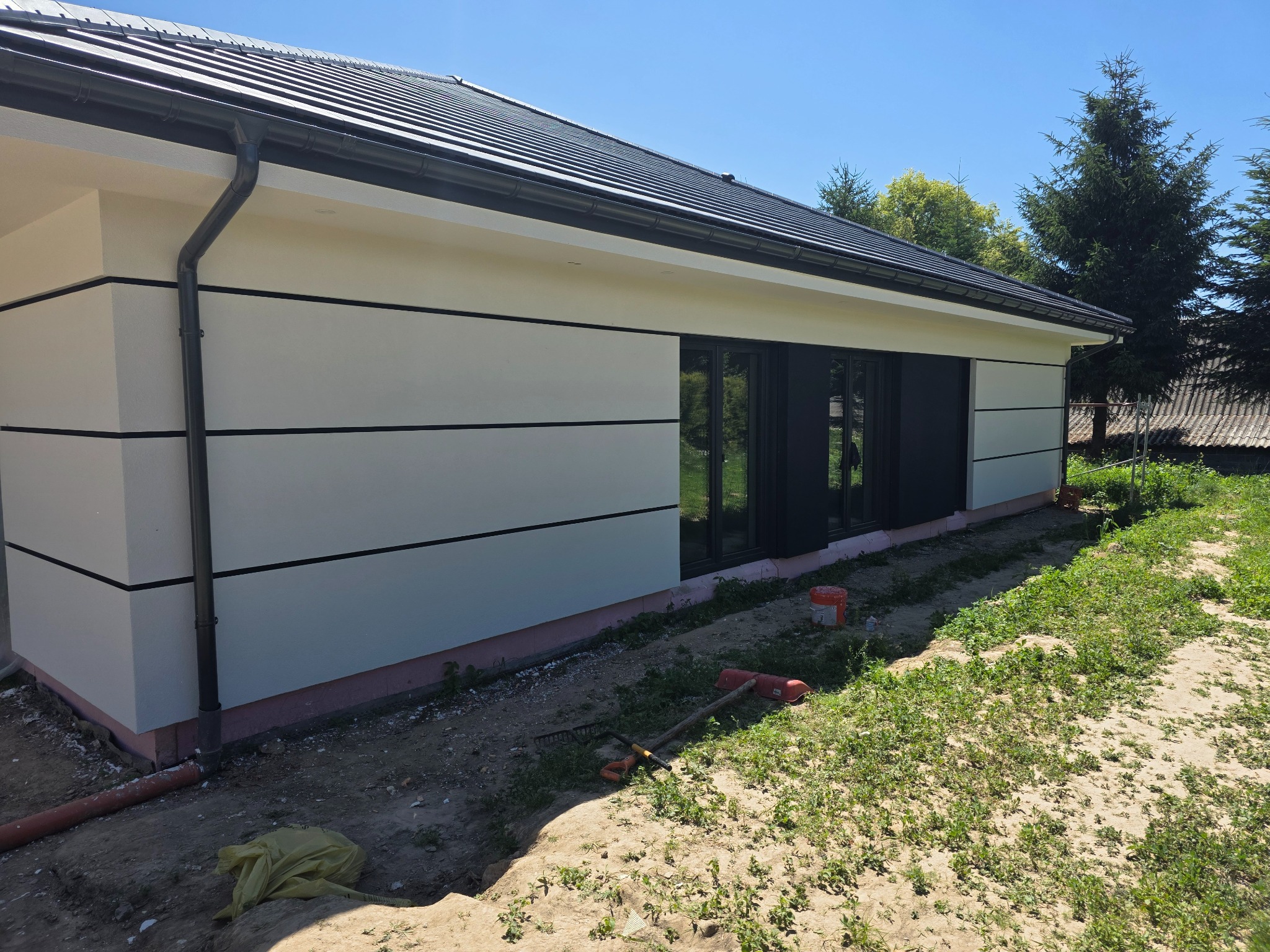 Elewacja domu jednorodzinnego z poziomymi, czarnymi pasami dekoracyjnymi. Widoczne okna, rynna i fragment dachu. W tle zieleń drzew i błękitne niebo.