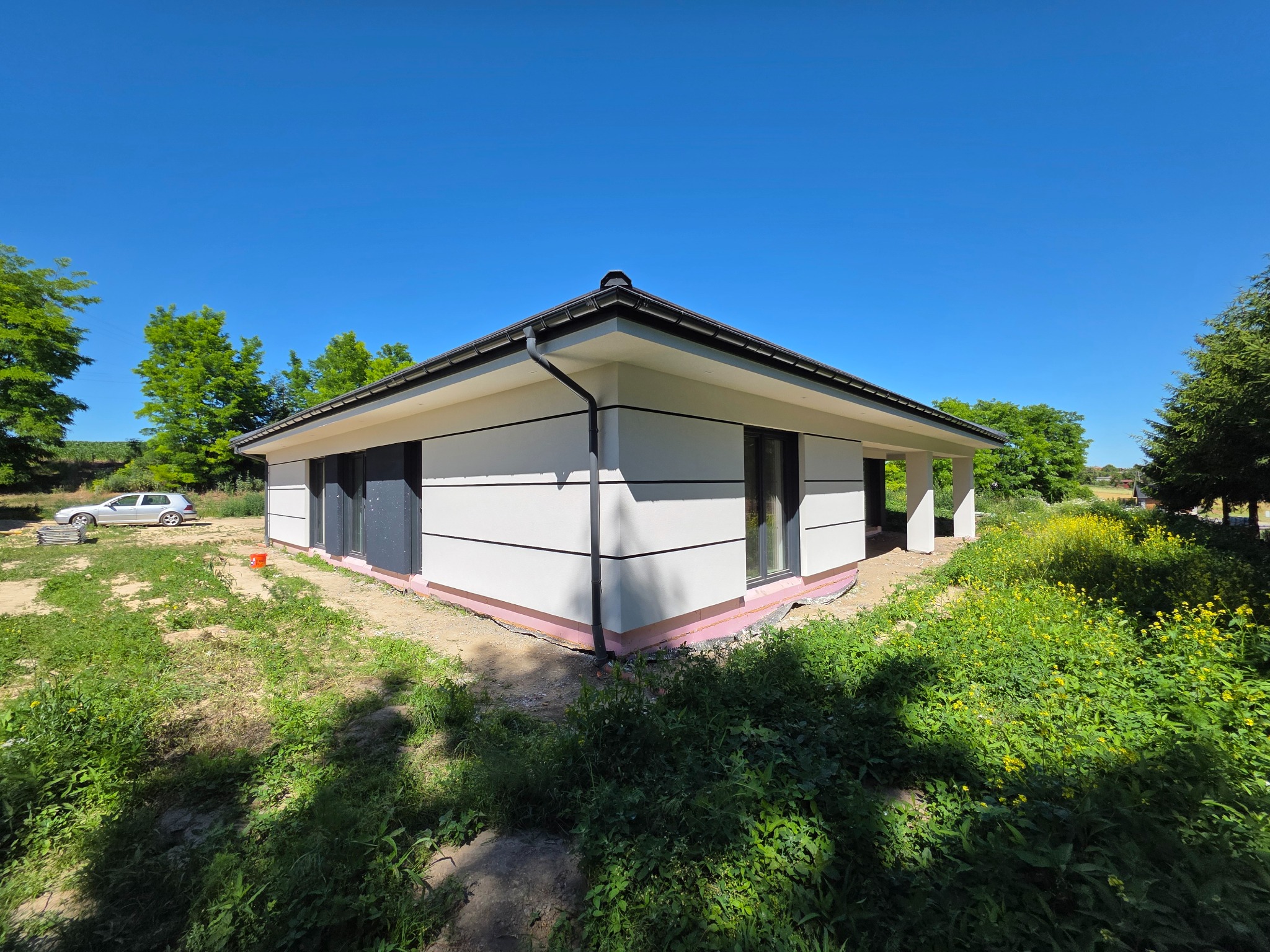 Nowoczesny, parterowy dom z białą elewacją i grafitowymi oknami, otoczony zielenią. Widoczna warstwa izolacji przy gruncie. Błękitne niebo w tle.