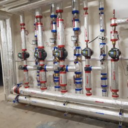 ARIONEX SPÓŁKA Z OGRANICZONĄ ODPOWIEDZIALNOŚCIĄ - Izolowane rury instalacji grzewczej z zaworami i licznikami ciepła, zamontowane wzdłuż ściany. Widoczne srebrne izolacje, czerwone i niebieskie elementy. W tle fragment ściany i otwór drzwiowy.