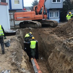 ARIONEX SPÓŁKA Z OGRANICZONĄ ODPOWIEDZIALNOŚCIĄ - Montaż rury kanalizacyjnej w wykopie przez pracowników w żółtych kamizelkach, z pomarańczową koparką Doosan w tle. Widoczne kontenery biurowe.