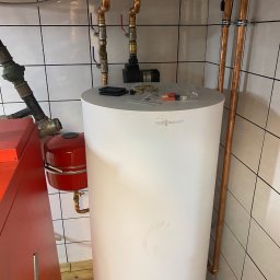 ARIONEX SPÓŁKA Z OGRANICZONĄ ODPOWIEDZIALNOŚCIĄ - Biały zasobnik ciepłej wody Viessmann z miedzianymi rurami i armaturą na tle białych płytek. Widoczny fragment czerwonego kotła. Instalacja w kotłowni.