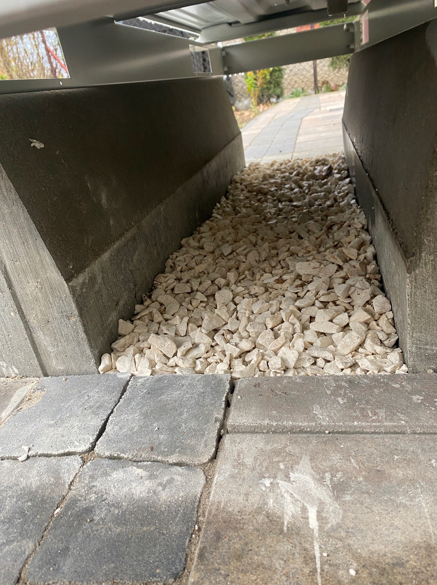 Drenaż z białego kruszywa w betonowym korycie, widok z dołu spod konstrukcji. Szara kostka brukowa w tle. Ujęcie z bliska, podkreślające detale.