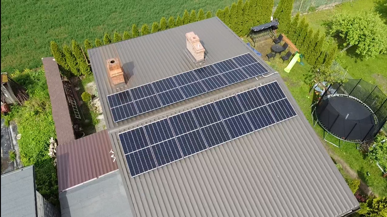 Widok z góry na panele fotowoltaiczne zamontowane na szarym dachu domu jednorodzinnego, obok komin z cegły, w tle zielony ogród z trampoliną.