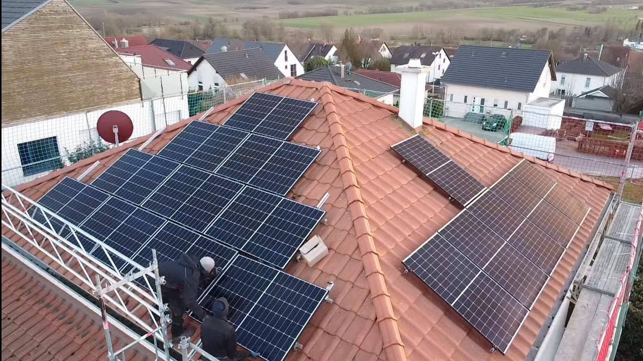 Montaż paneli fotowoltaicznych na dachu z czerwonej dachówki. Widoczni pracownicy na rusztowaniu, instalujący panele. W tle zabudowa jednorodzinna i pola.