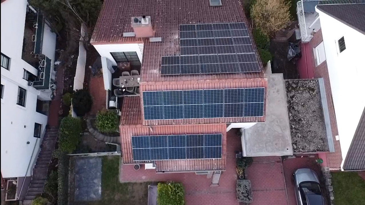 Widok z drona na dom z czerwoną dachówką i panelami fotowoltaicznymi. Panele rozmieszczone na różnych poziomach dachu. Widoczne otoczenie domu: ogród, sąsiednie budynki.