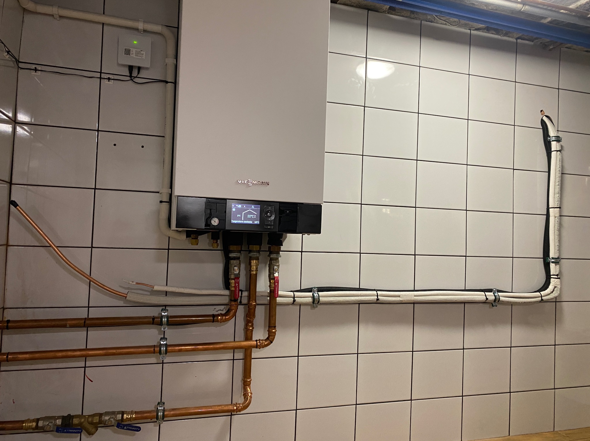 Nowoczesny, wiszący piec gazowy Viessmann z miedzianą instalacją, zamontowany na ścianie z białych płytek. Widoczne rury i przewody.