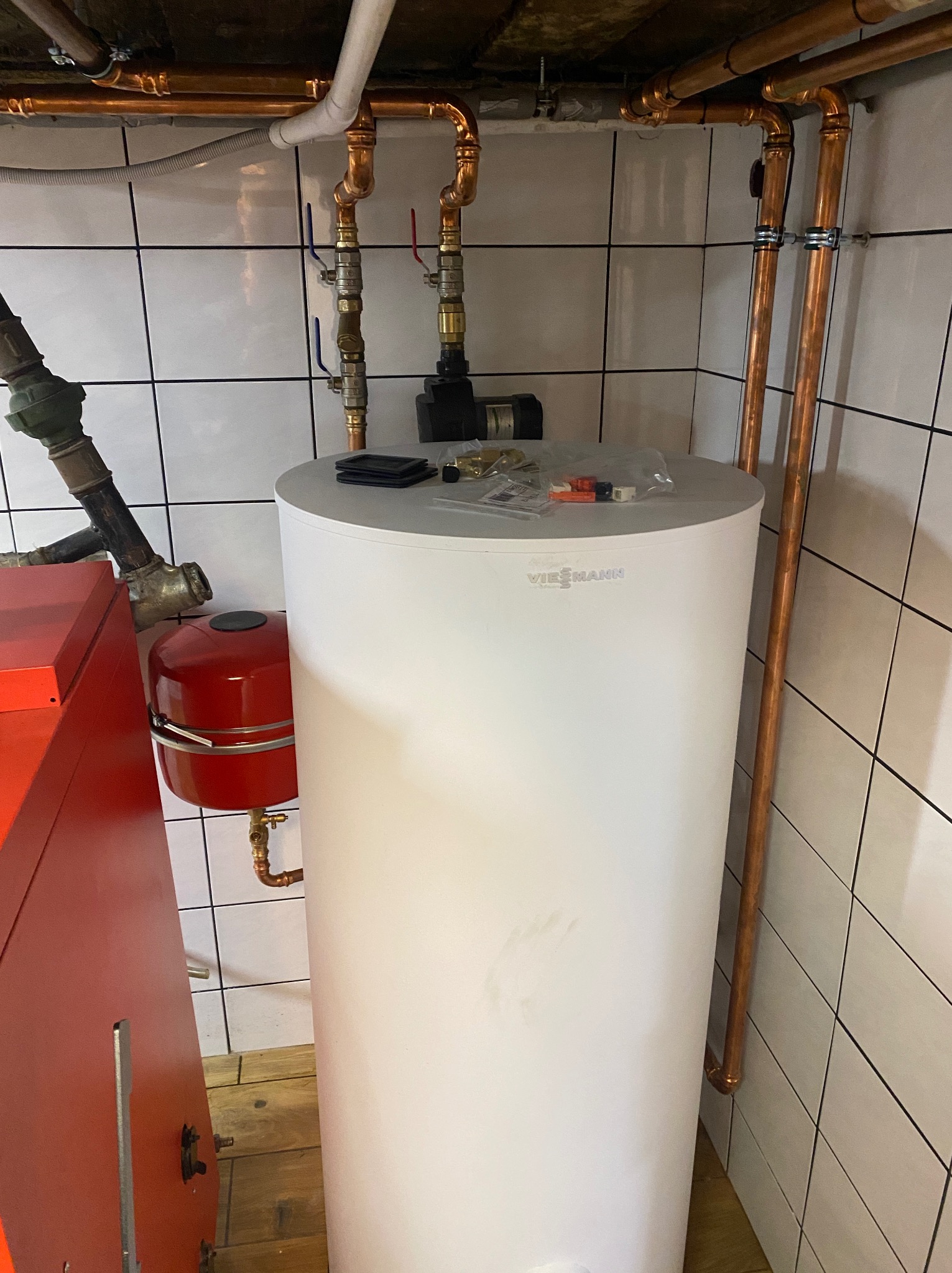 Biały zasobnik ciepłej wody Viessmann z miedzianymi rurami i armaturą na tle białych płytek. Widoczny fragment czerwonego kotła. Instalacja w kotłowni.