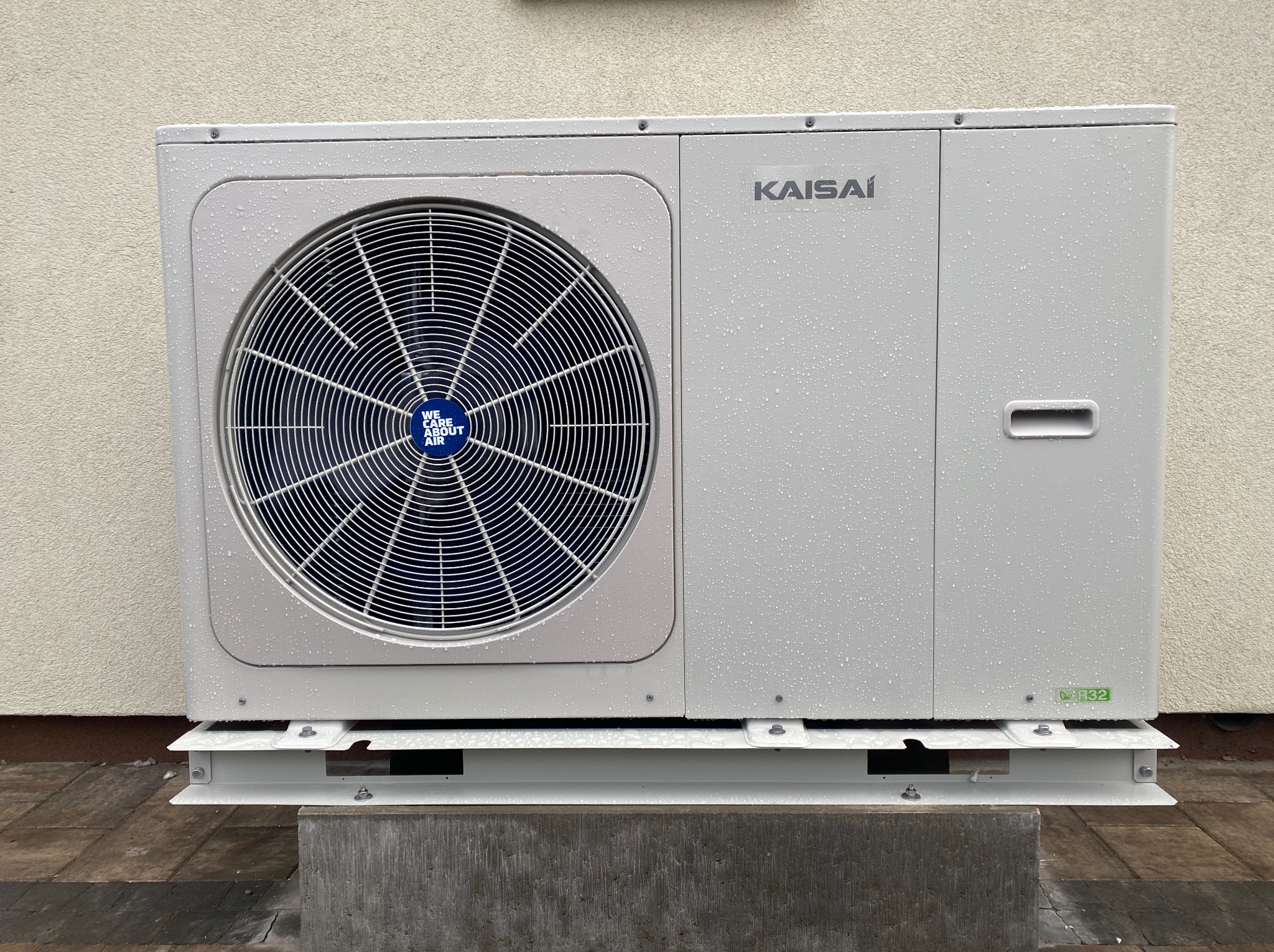 Biała jednostka zewnętrzna klimatyzacji Kaisai, zamontowana na metalowej podstawie, z widocznymi kroplami wody na obudowie. Tło stanowi jasna ściana budynku.