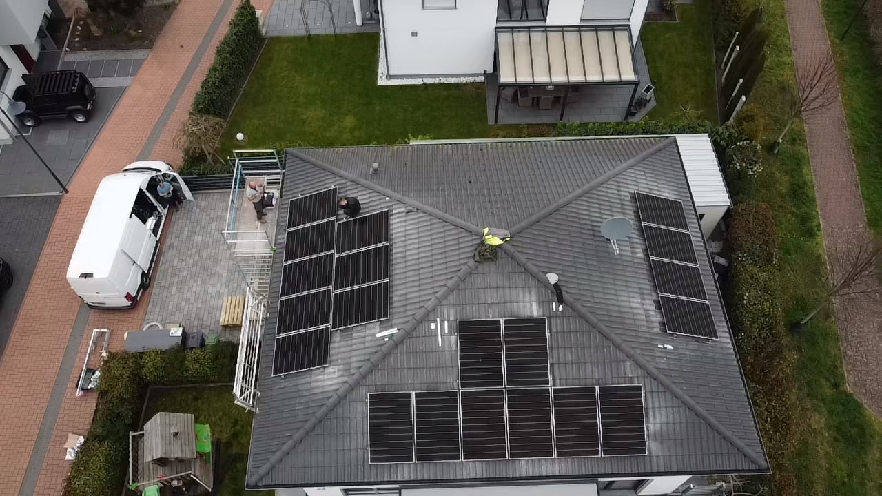 Montaż paneli fotowoltaicznych na dachu domu jednorodzinnego, widok z góry. Pracownicy w kamizelkach odblaskowych, biały bus i rusztowanie obok budynku.