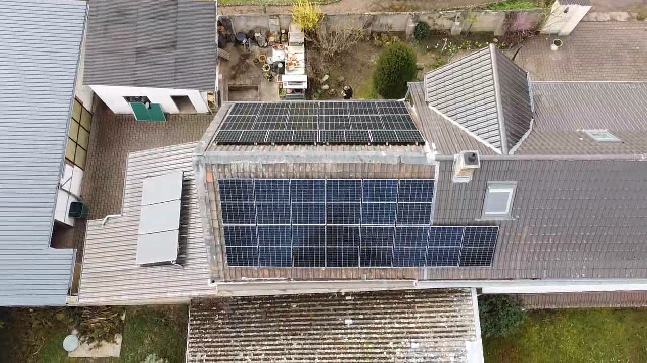 Widok z góry na dom z panelami fotowoltaicznymi na dachu. Panele ułożone w kilku sekcjach, na różnych poziomach dachu. Widoczne otoczenie domu i inne budynki.