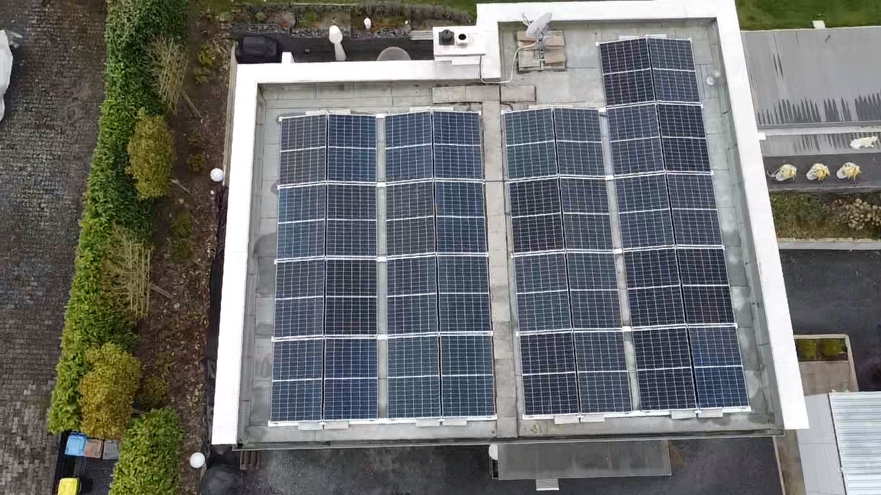 Widok z góry na panele fotowoltaiczne na płaskim dachu budynku z anteną satelitarną. Obok dachu widoczna zieleń i fragmenty innych budynków.