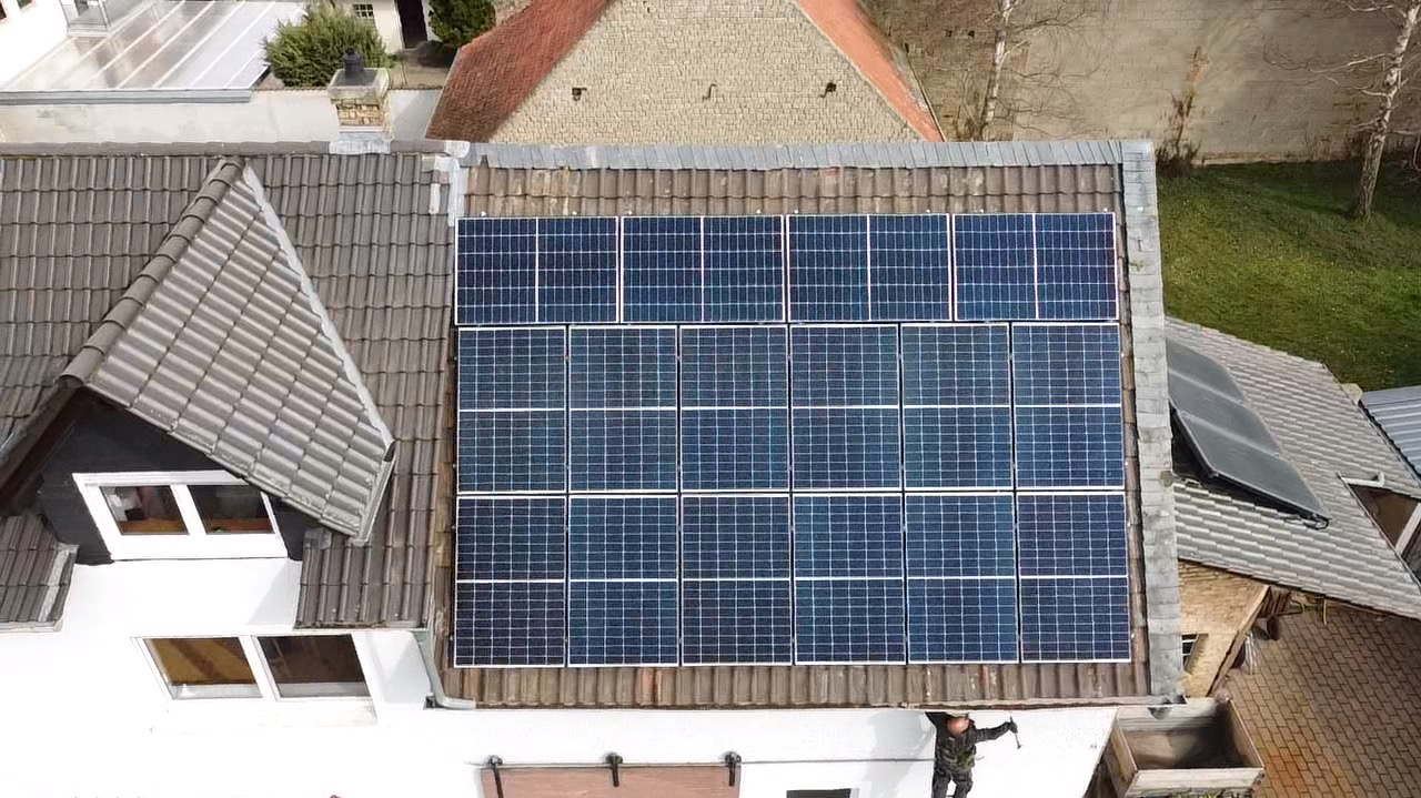 Widok z góry na instalację fotowoltaiczną na dachu domu z szarymi dachówkami. Na dole kadru widoczna osoba w kombinezonie montażowym.