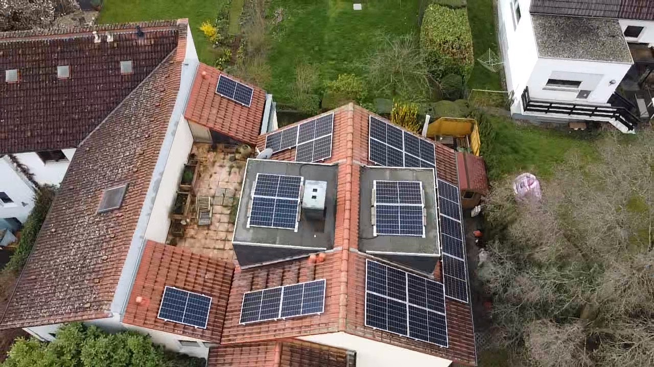 Widok z góry na dom z panelami fotowoltaicznymi na dachu, otoczony zielenią. Panele różnej wielkości, rozmieszczone na różnych poziomach dachu. Widoczne są sąsiednie budynki.