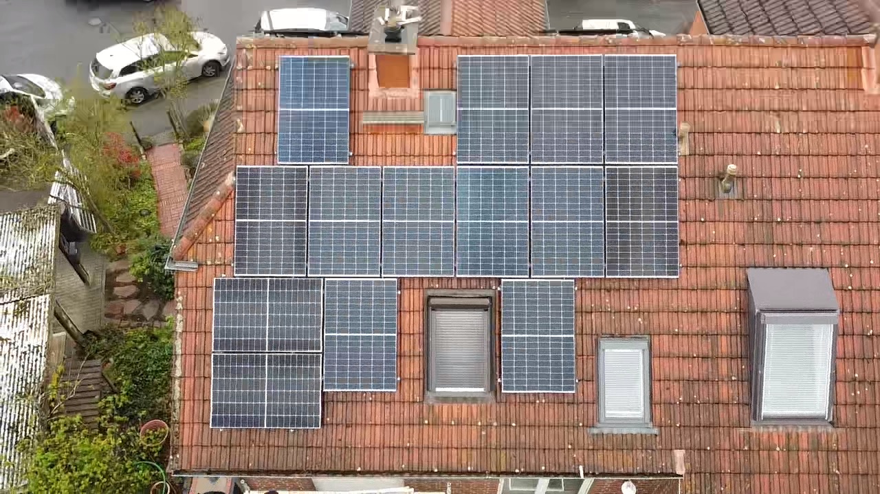 Widok z góry na panele fotowoltaiczne na dachu z czerwonej dachówki, z oknami dachowymi i fragmentami zieleni. Układ paneli asymetryczny, z widocznymi elementami montażowymi.