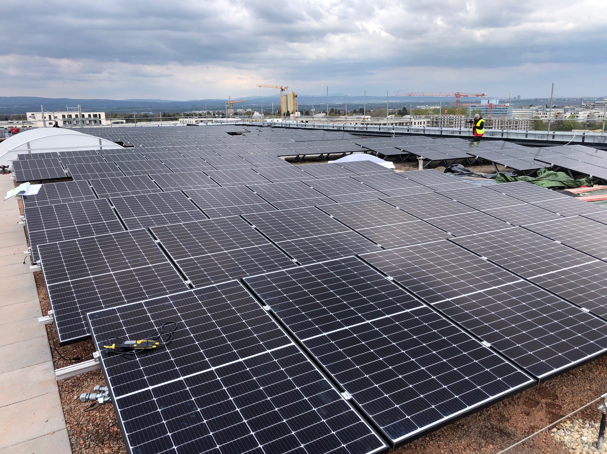 Rozległa instalacja fotowoltaiczna na dachu budynku, z widoczną panoramą miasta w tle i pracownikiem w odblaskowej kamizelce. Widoczne narzędzia i elementy montażowe.
