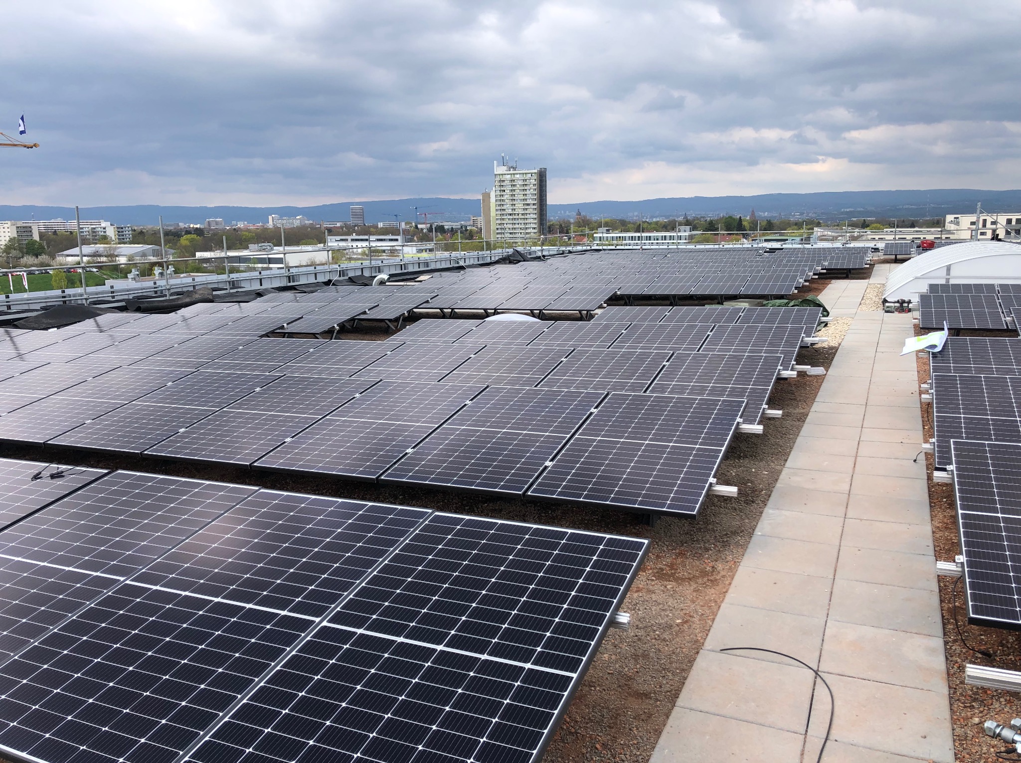Rozległa instalacja fotowoltaiczna na dachu budynku, widok z góry. Panele ułożone rzędami, w tle zabudowa miejska i pochmurne niebo. Widoczne elementy konstrukcyjne i kable.