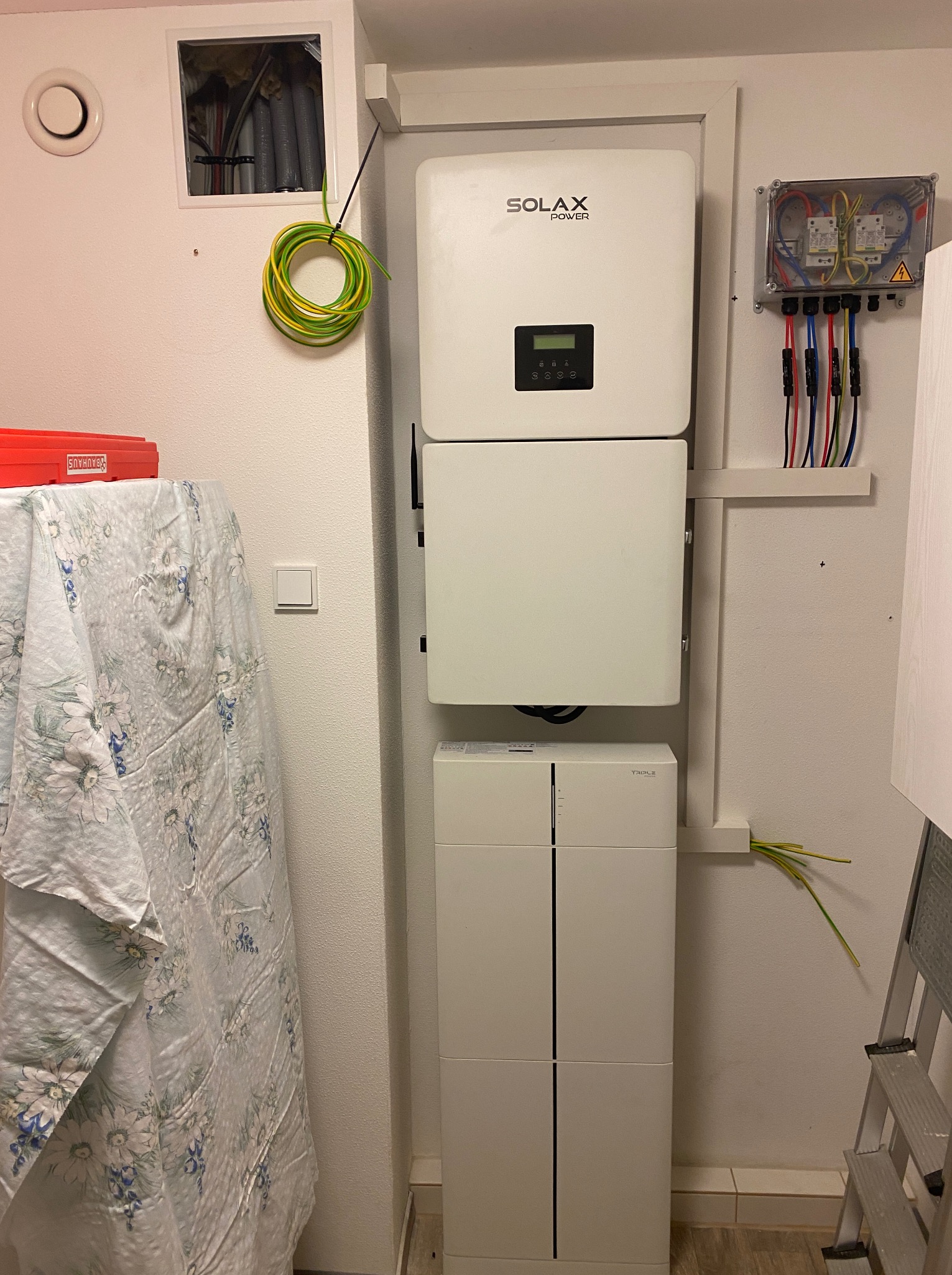 Biały inwerter Solax Power z baterią i zabezpieczeniami na ścianie, widoczne kable. Instalacja fotowoltaiczna w pomieszczeniu, drabina oparta o ścianę.