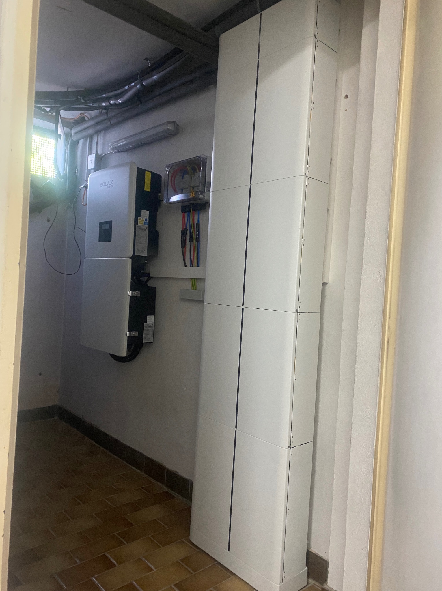 Biały inwerter fotowoltaiczny Solax Power zamontowany na ścianie w piwnicy, obok pionowa, biała obudowa akumulatorów. Widoczne okablowanie i skrzynka elektryczna.