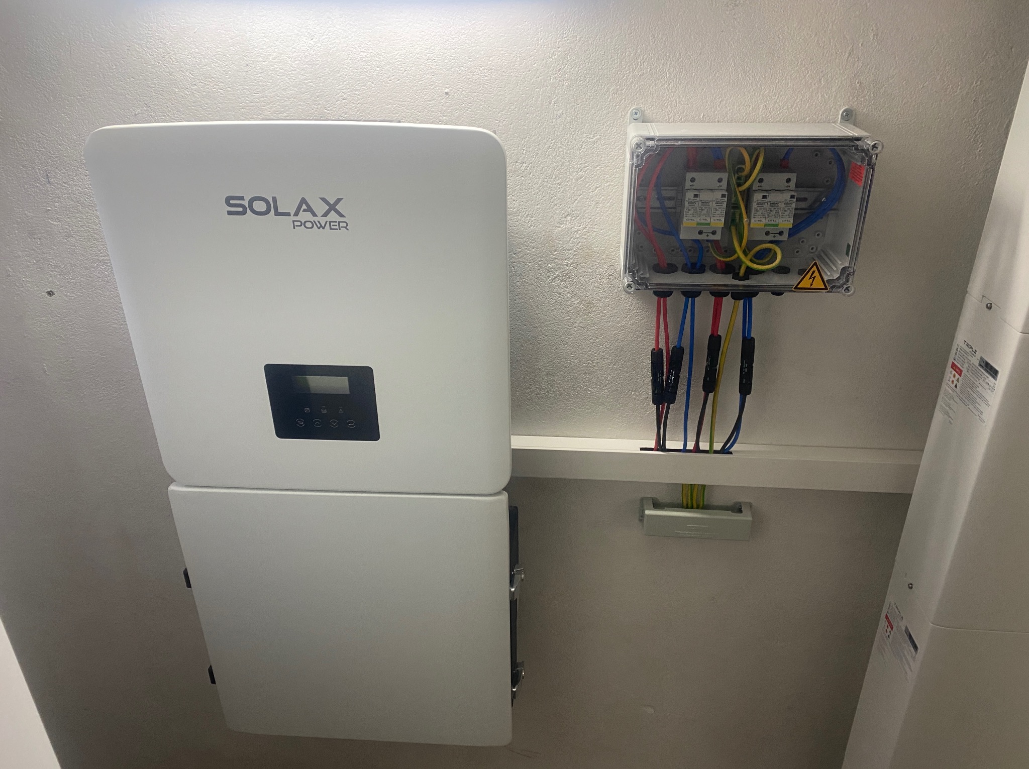 Biały inwerter Solax Power zamontowany na ścianie obok skrzynki z zabezpieczeniami, widoczne przewody elektryczne. Instalacja fotowoltaiczna wewnątrz budynku.