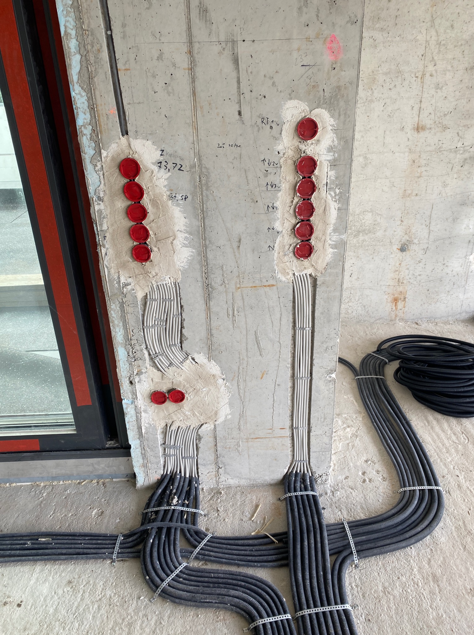 Montaż przewodów elektrycznych w ścianie betonowej, zabezpieczonych czerwonymi zaślepkami i przymocowanych metalowymi klamrami. Widoczne szare i czarne rury instalacyjne.