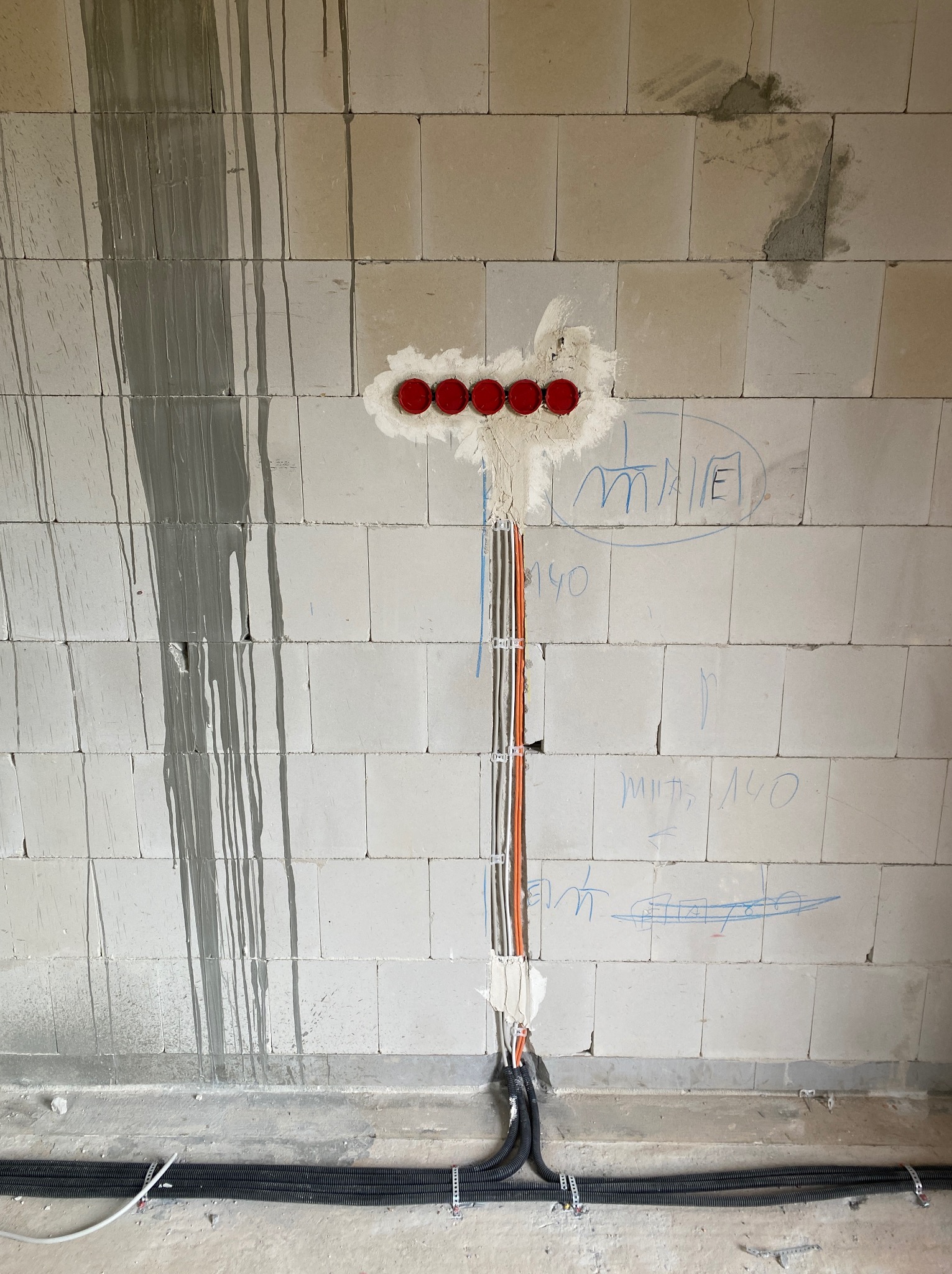 Instalacja elektryczna w surowym murze z betonu komórkowego. Widoczne kable w peszlach i puszki pod gniazdka z czerwonymi zaślepkami. Ślady po ołówku na ścianie.