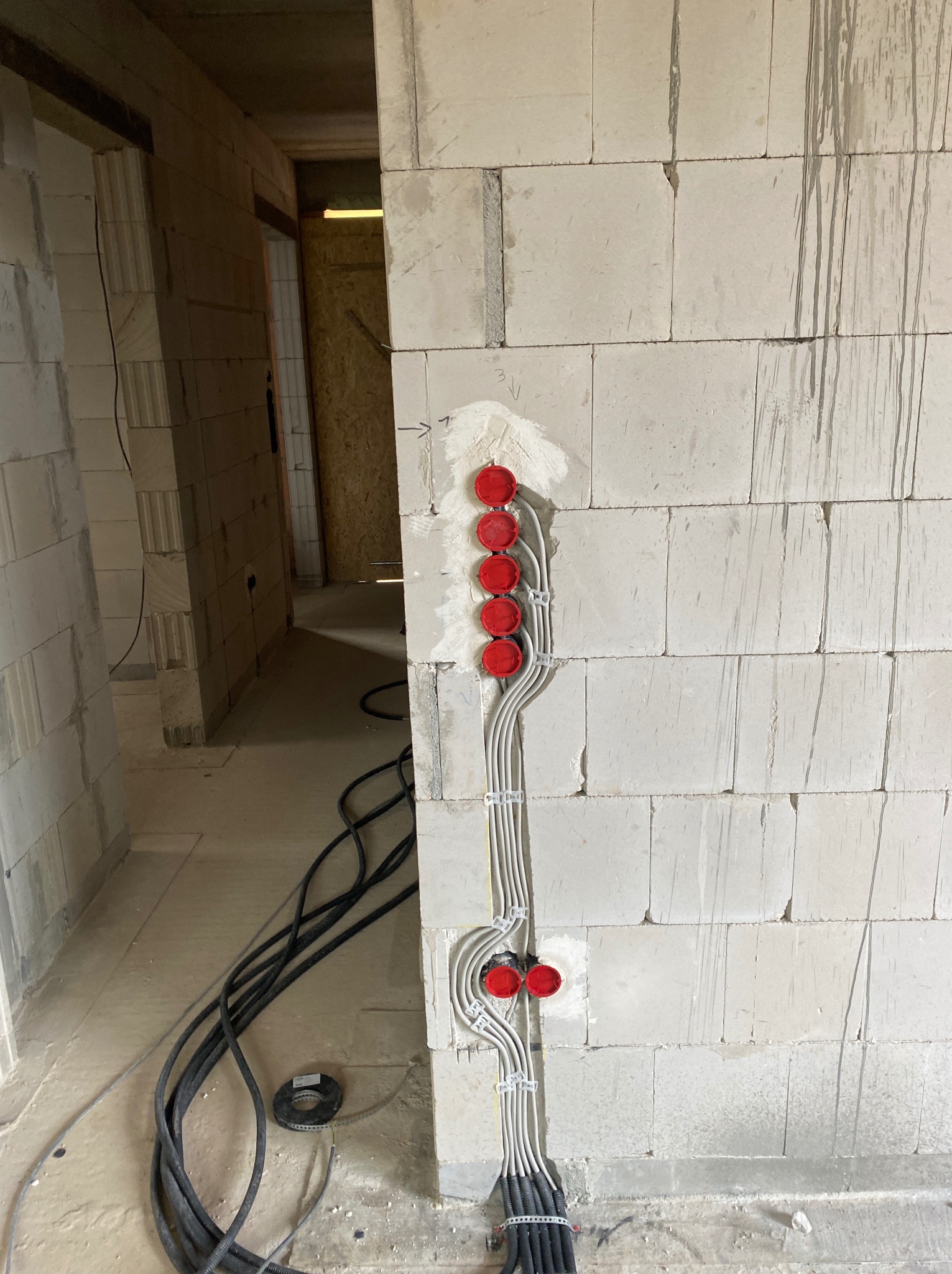 Ściana z instalacją elektryczną w trakcie montażu: widoczne przewody w peszlach i czerwone puszki pod gniazdka. Surowy stan, szare ściany z betonu komórkowego.