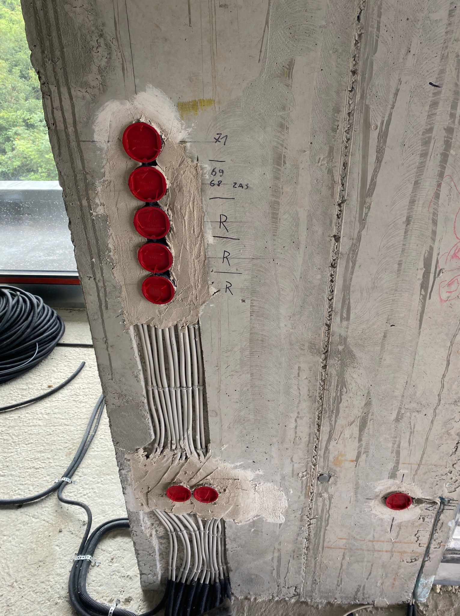 Betonowy słup z instalacją elektryczną w trakcie montażu: czerwone puszki i białe przewody w bruzdach. Widoczne oznaczenia i kable w peszlach. Surowy stan wykończenia.