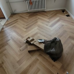 Konrad Pfeiffer - Ułożony parkiet w jodełkę francuską, widoczne resztki materiałów i narzędzia po pracy. Jasne panele, grzejnik pod oknem, fragment podłogi bez listew przypodłogowych.