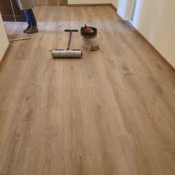 Konrad Pfeiffer - Jasne panele podłogowe z widocznym wałkiem dociskowym i pojemnikiem z narzędziami. Fragment osoby w kadrze. Widoczny grzejnik na ścianie.