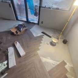 Konrad Pfeiffer - Układanie paneli w jodełkę, widoczne narzędzia i materiały budowlane, folia ochronna na oknach. Częściowo ułożona podłoga, lampa budowlana.