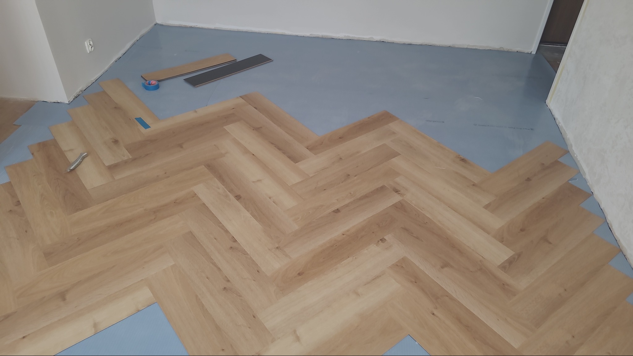 Fragment podłogi z ułożonym parkietem w jodełkę, widoczne narzędzia i taśma malarska. Podkład pod panele w kolorze szarym. Częściowo wykończone pomieszczenie.