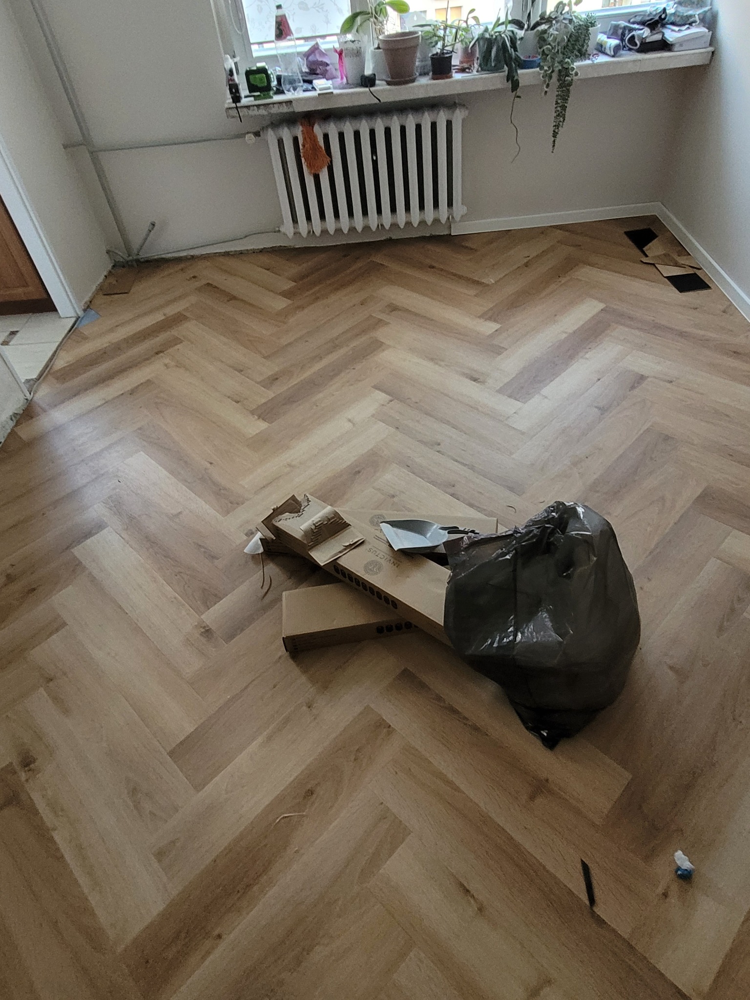 Ułożony parkiet w jodełkę francuską, widoczne resztki materiałów i narzędzia po pracy. Jasne panele, grzejnik pod oknem, fragment podłogi bez listew przypodłogowych.