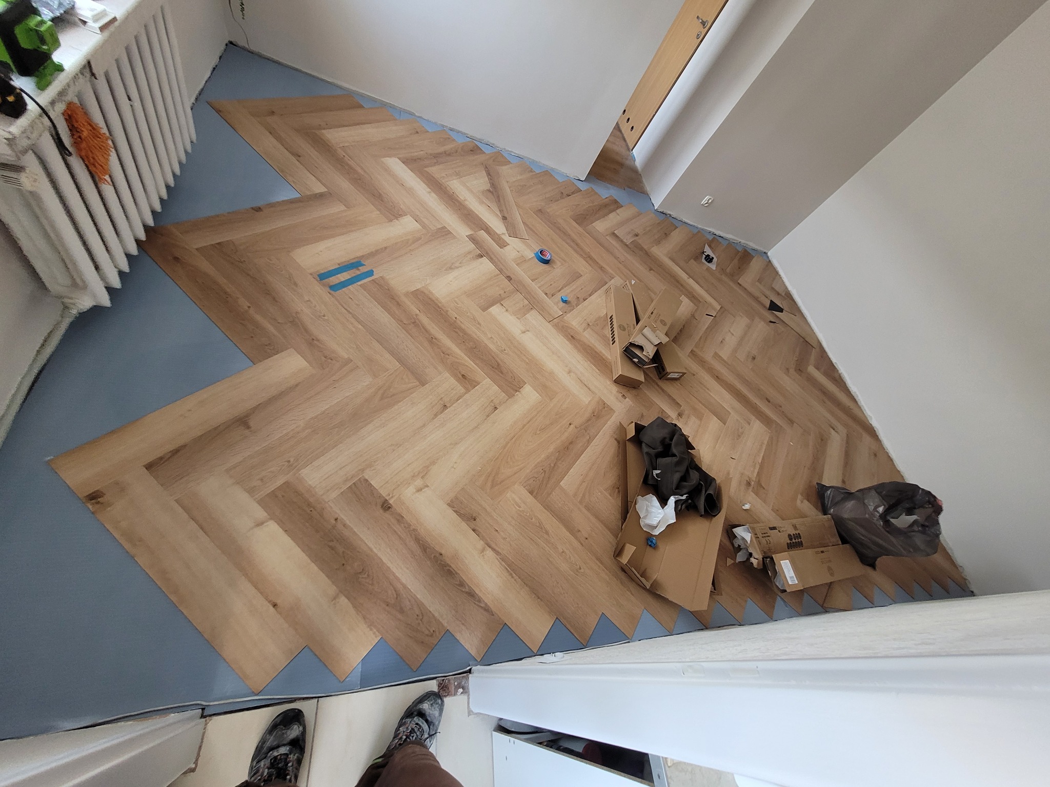 Układanie paneli w jodełkę na podkładzie. Widoczne kartony po panelach, resztki materiałów. Częściowo ułożona podłoga w jasnym odcieniu drewna.