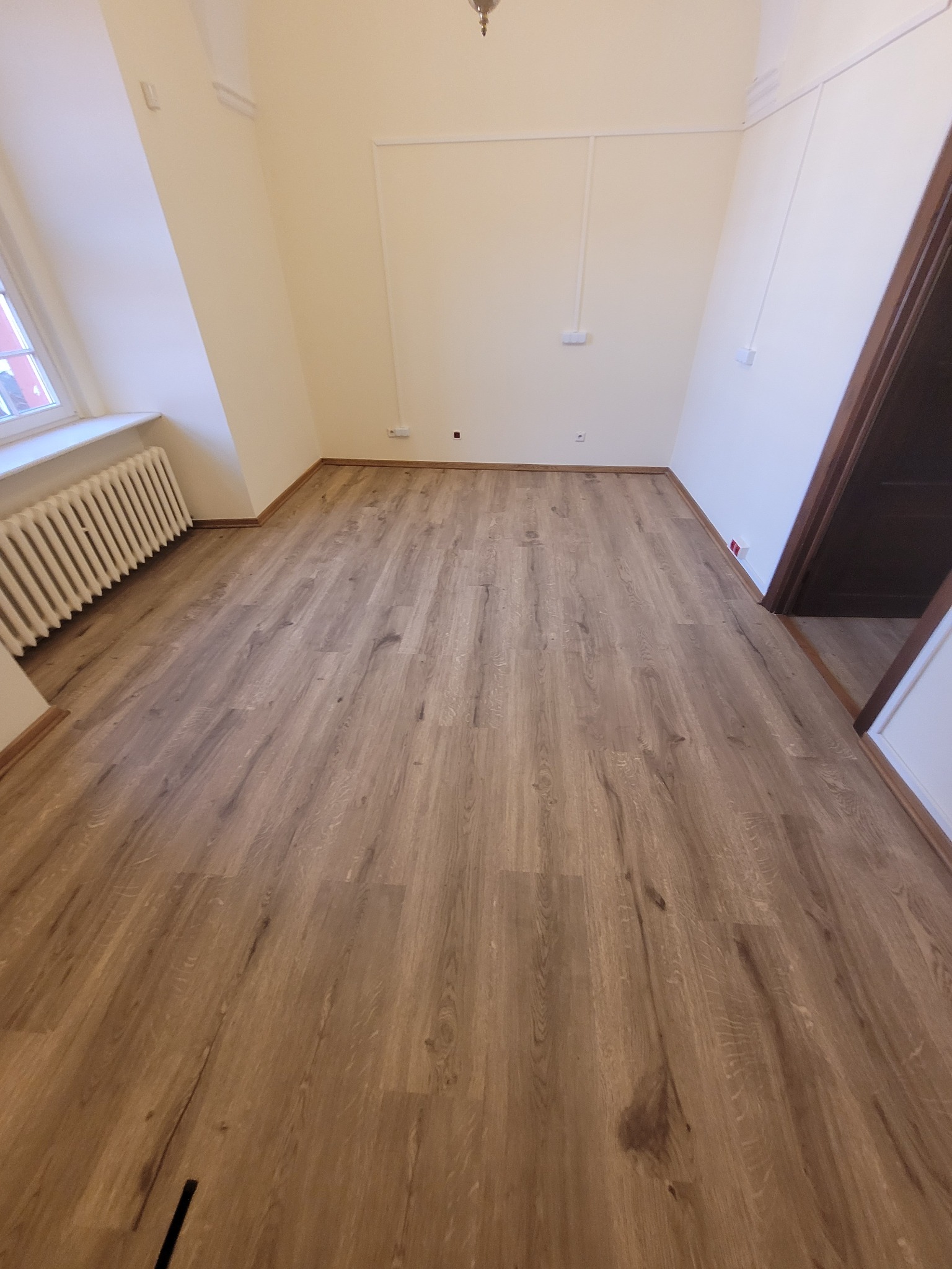 Jasne panele podłogowe w pokoju z grzejnikiem i oknem. Widoczne listwy przypodłogowe i fragment drzwi. Ujęcie z niskiej perspektywy.