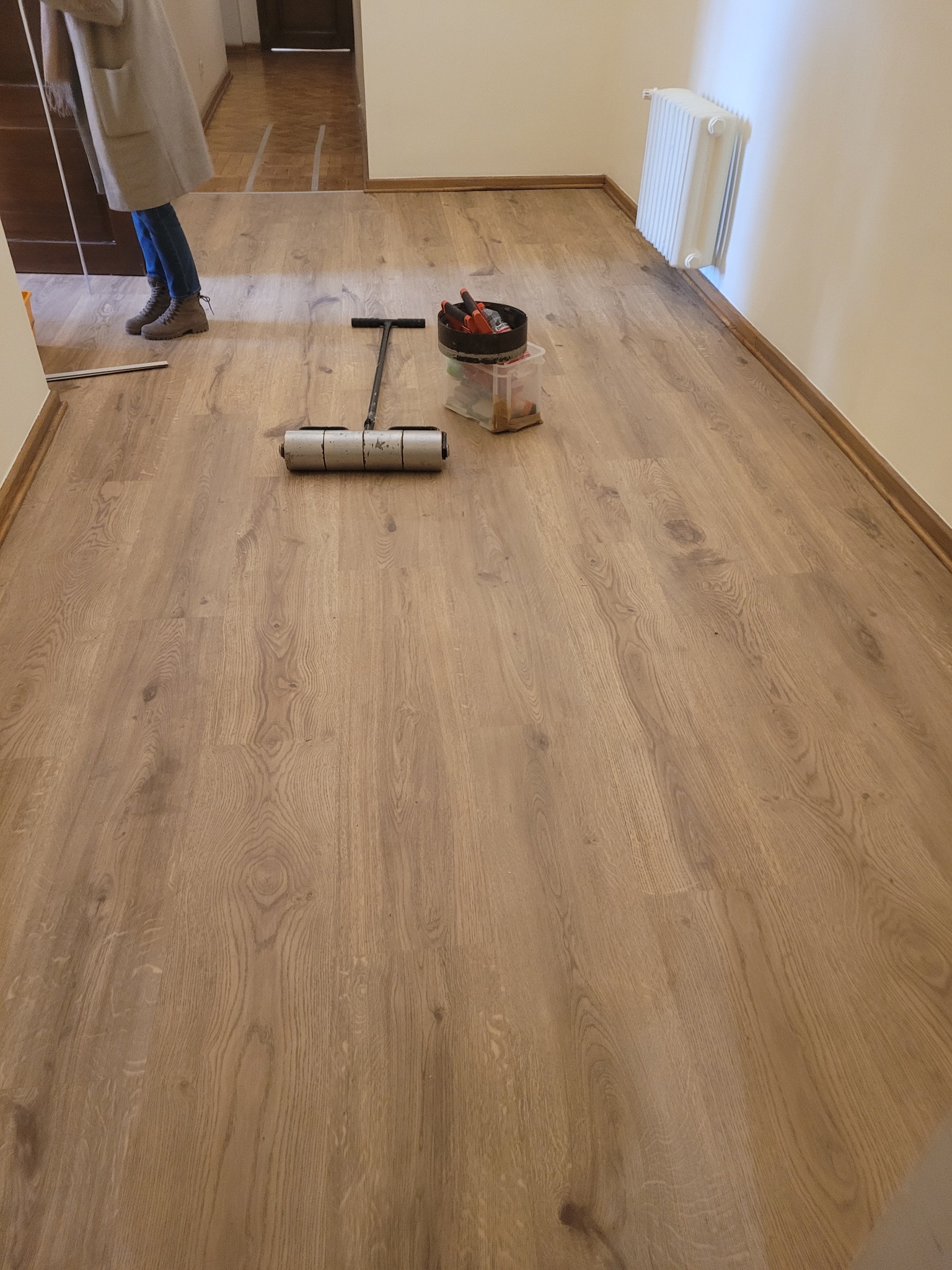 Jasne panele podłogowe z widocznym wałkiem dociskowym i pojemnikiem z narzędziami. Fragment osoby w kadrze. Widoczny grzejnik na ścianie.