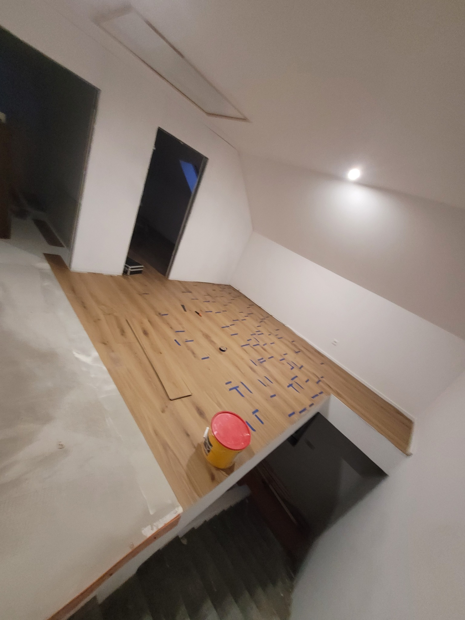 Poddasze w trakcie prac: panele podłogowe z niebieskimi znacznikami, wiadro farby. Widoczne skosy i drzwi. Częściowo ułożona podłoga.