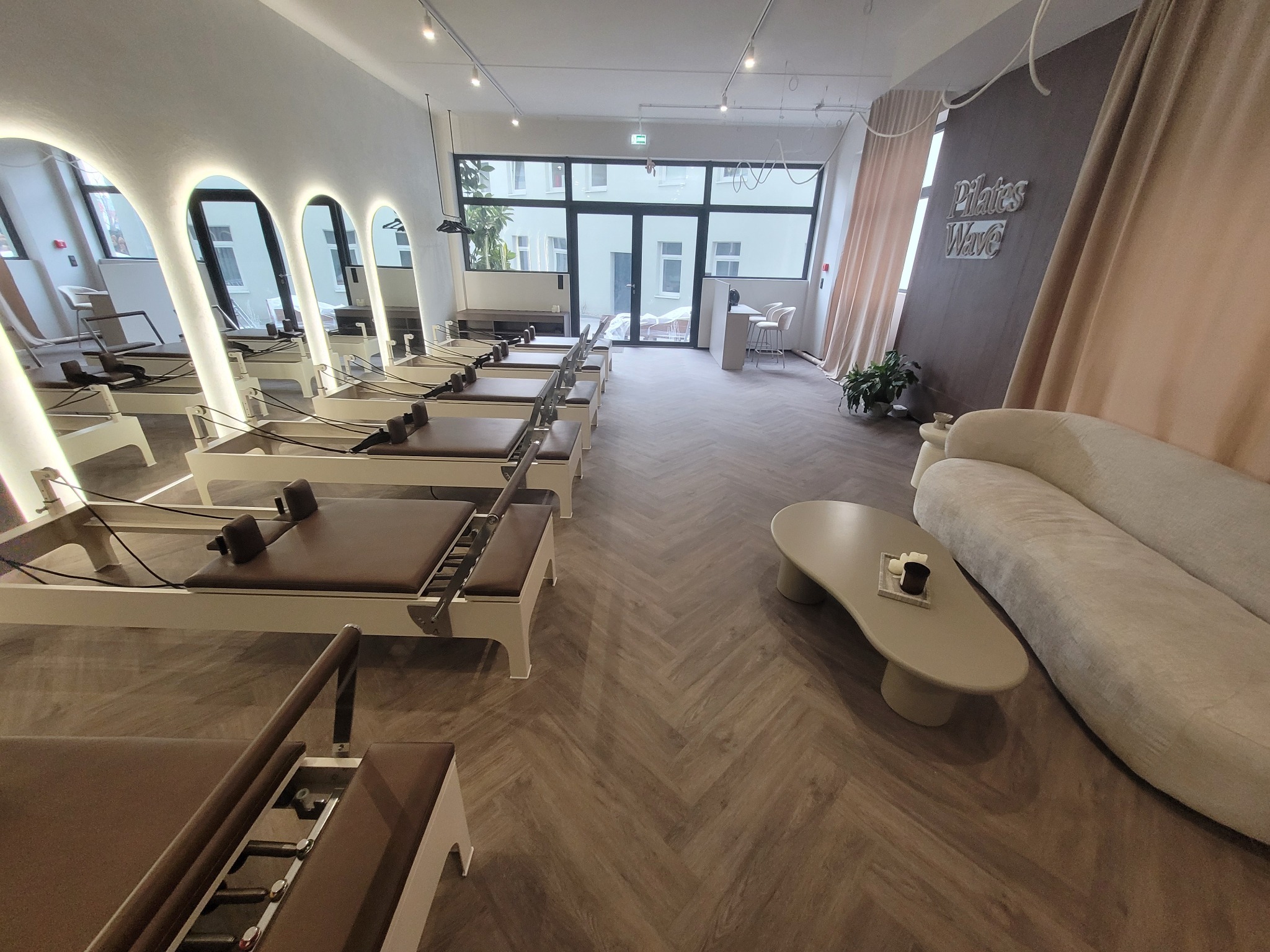 Jasne studio pilates z parkietem w jodełkę, rzędem maszyn do ćwiczeń i minimalistycznym wystrojem. Duże okna wpuszczają naturalne światło.