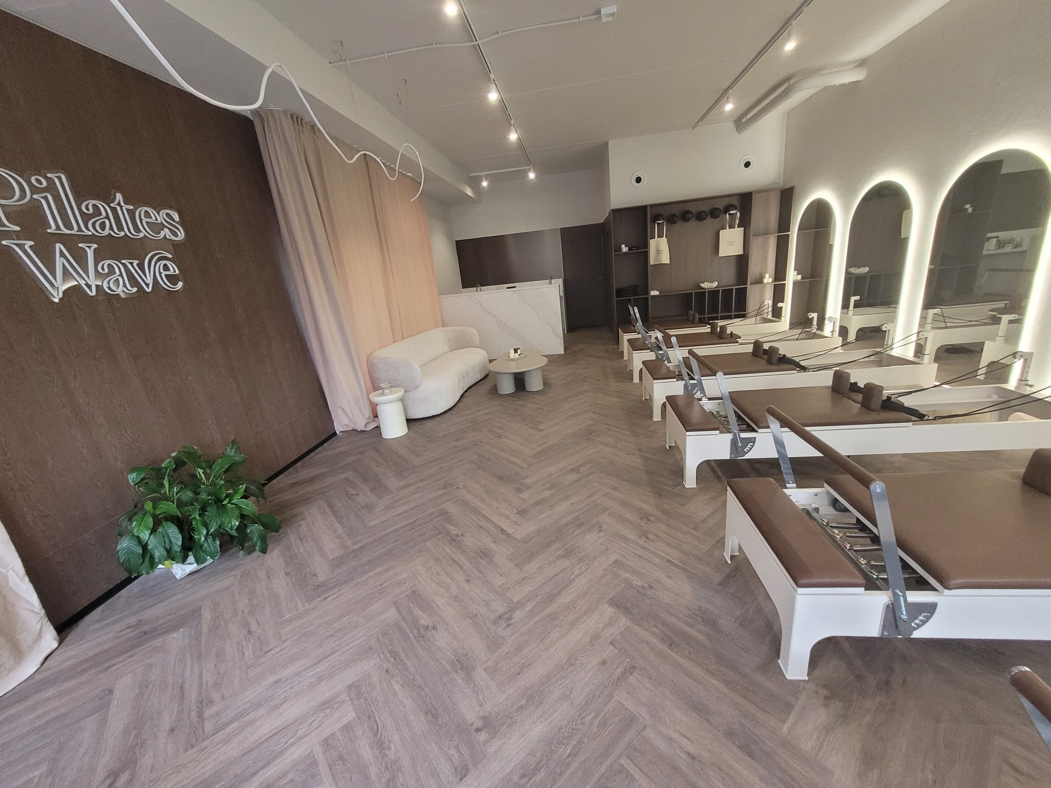 Jasne, przestronne studio pilates z parkietem ułożonym we wzór jodełki, nowoczesnym oświetleniem i sprzętem do ćwiczeń. Minimalistyczny design z akcentami drewna i zieleni.