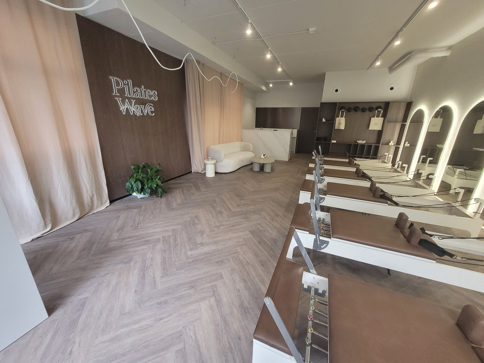 Jasna podłoga ułożona w jodełkę w przestronnym wnętrzu studia Pilates Wave. W tle biała sofa i sprzęt do ćwiczeń. Minimalistyczny design i naturalne światło.