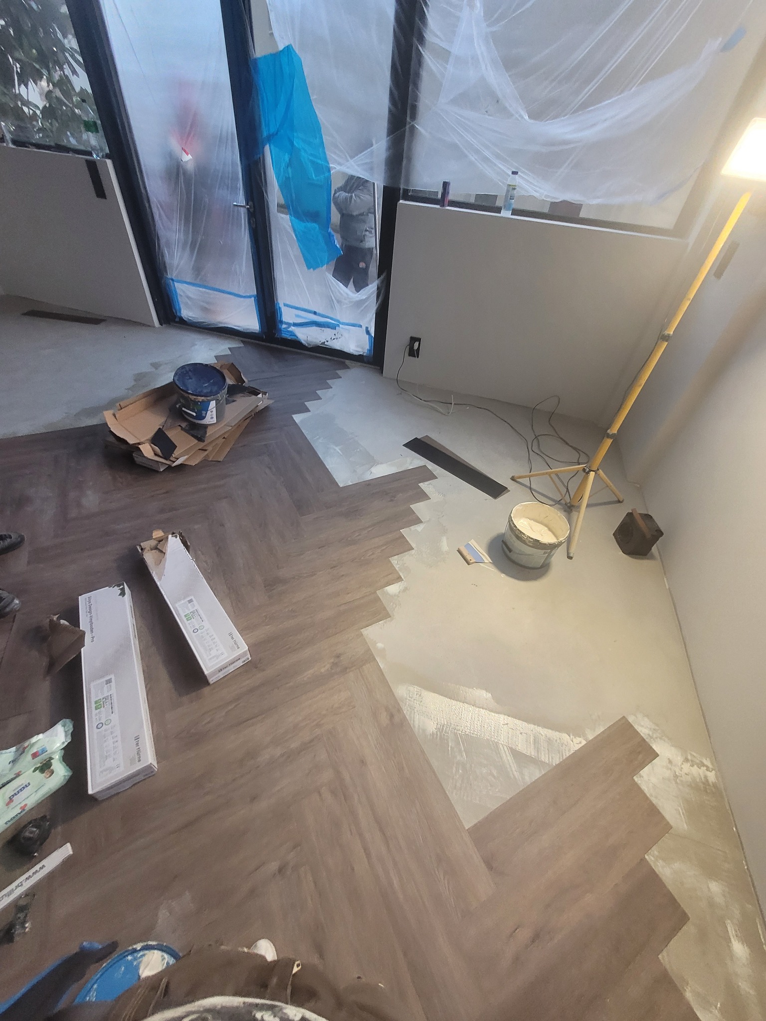 Układanie paneli w jodełkę, widoczne narzędzia i materiały budowlane, folia ochronna na oknach. Częściowo ułożona podłoga, lampa budowlana.