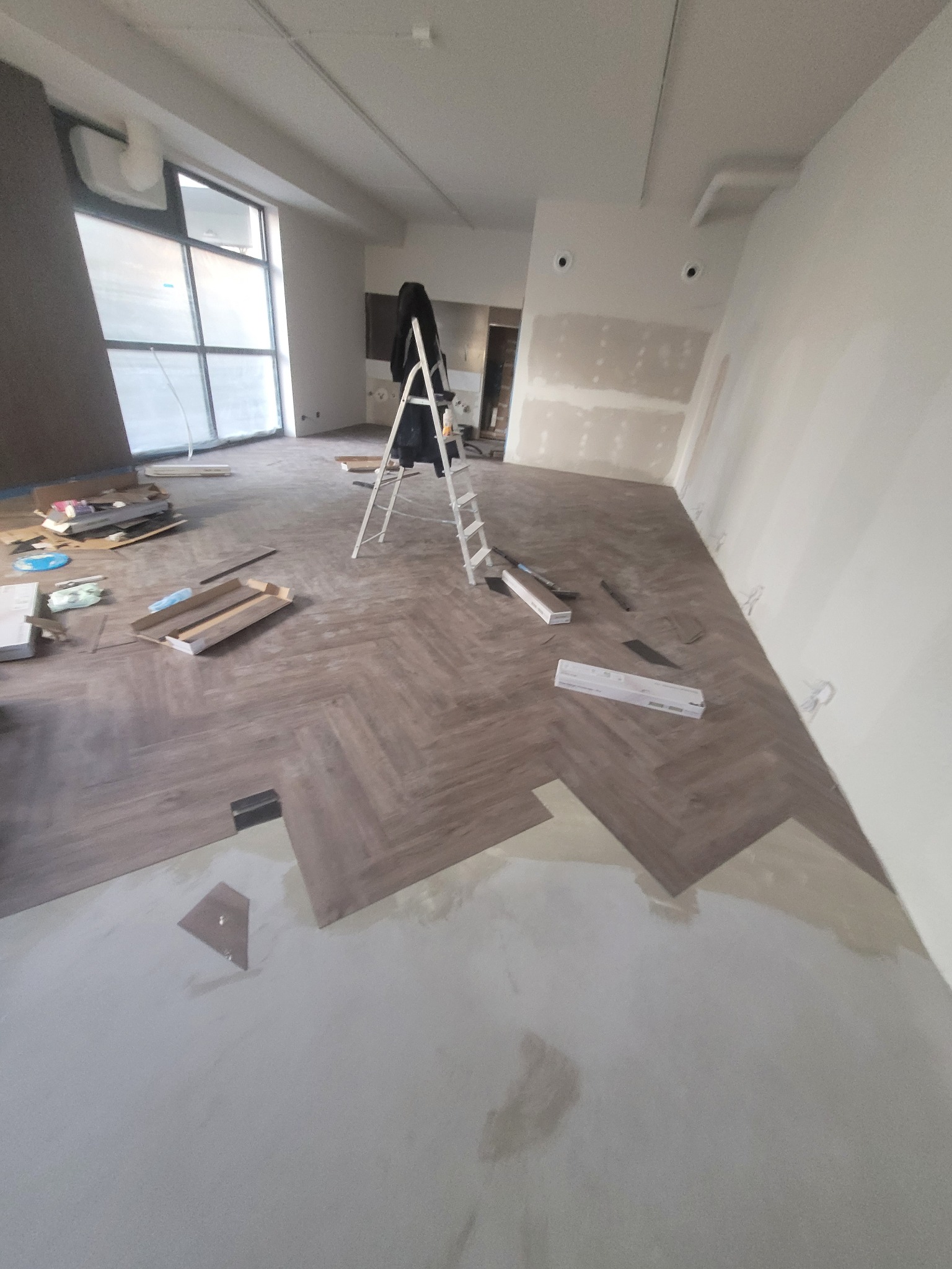 Układanie paneli w jodełkę w jasnym wnętrzu. Widoczny fragment podkładu, rozłożone panele i drabina. Duże okno z lewej strony. Etap prac wykończeniowych.