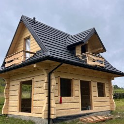 BaliHouse - Surowa drewniana konstrukcja domu z czarnym dachem, widoczne niezabudowane otwory okienne i prowizoryczne barierki balkonowe. Dom w trakcie budowy na tle zieleni.