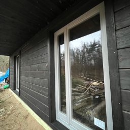 BaliHouse - Ciemna, drewniana elewacja domu z białymi oknami. Widoczne deski elewacyjne i fragment terenu budowy. W oknach odbija się las.