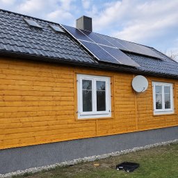 BaliHouse - Dom z drewnianą elewacją w odcieniach brązu, czarnym dachem z panelami słonecznymi i białymi oknami. Widoczna antena satelitarna i fragment ogrodu.