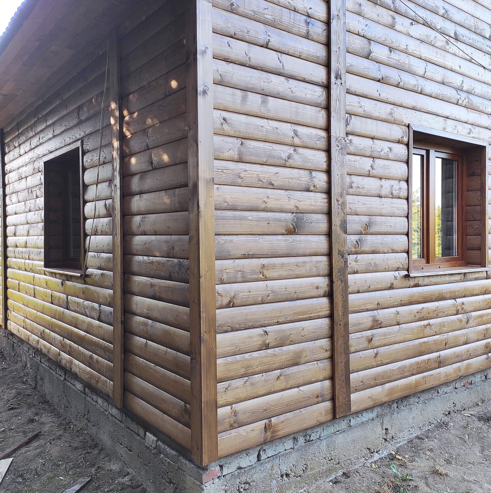 Fragment ściany domu z bali, z widocznymi oknami i fundamentem. Drewno pomalowane na brązowo, widoczne słoje. Ujęcie z dołu, podkreślające solidność konstrukcji.