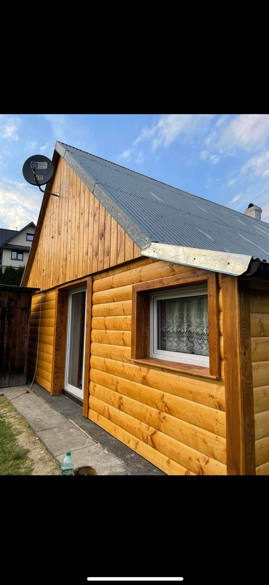 Drewniana elewacja domu z metalowym dachem i anteną satelitarną. Widoczne okno z firanką i fragment chodnika z butelką wody. Jasne drewno, szary dach.