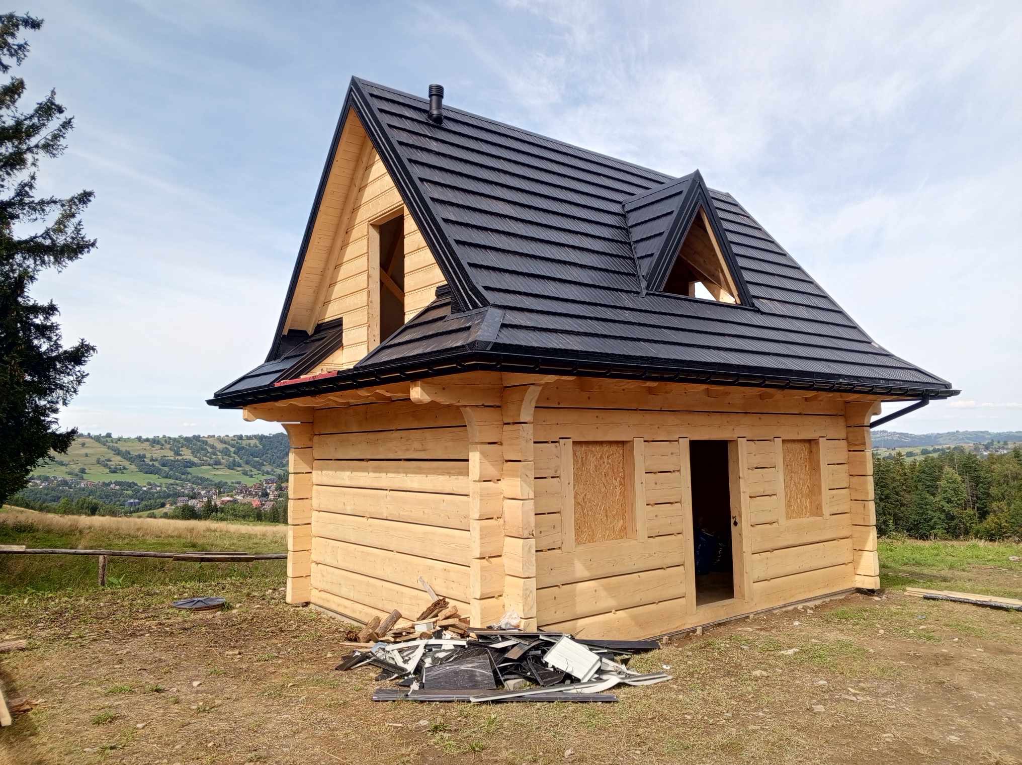 Surowa drewniana konstrukcja domu z czarnym dachem na wzgórzu, widok na dolinę w tle. Częściowo zabite okna, resztki materiałów budowlanych przed budynkiem.