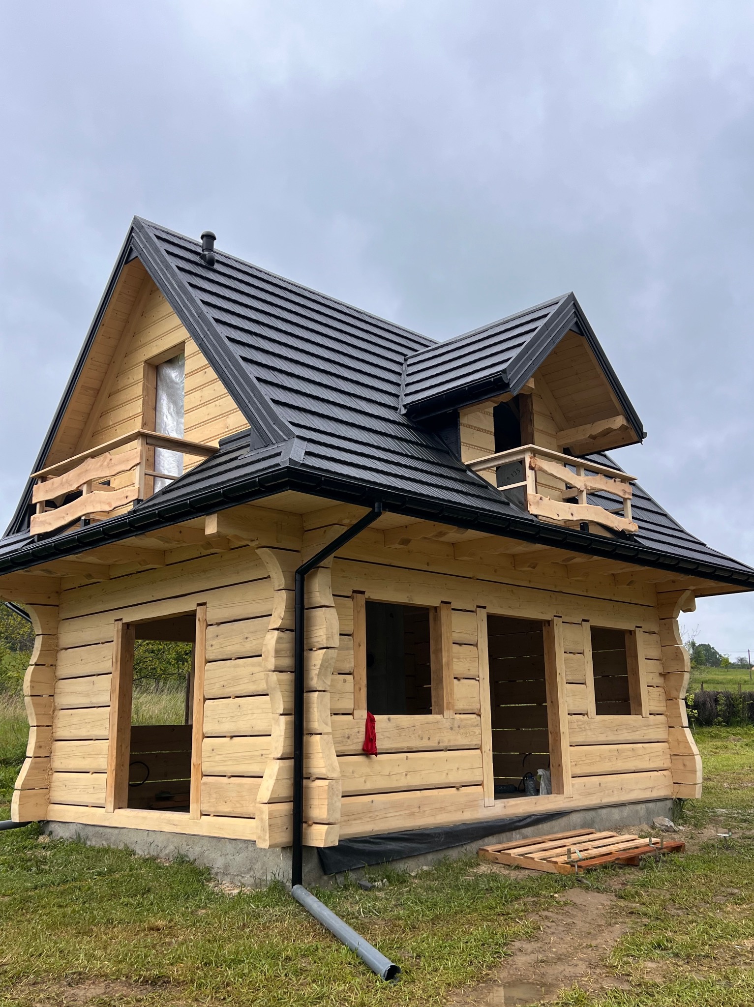 Surowa drewniana konstrukcja domu z czarnym dachem, widoczne niezabudowane otwory okienne i prowizoryczne barierki balkonowe. Dom w trakcie budowy na tle zieleni.