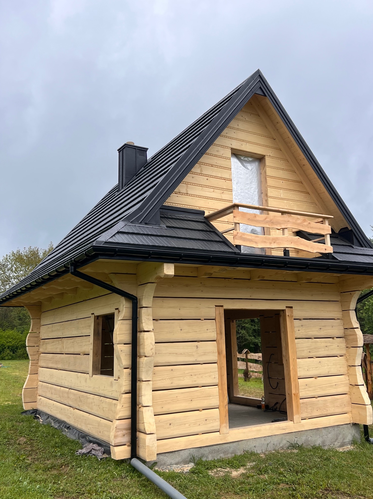 Surowa bryła domu z bali z czarnym dachem i niedokończoną balustradą balkonową. Widoczne detale obróbki drewna i brakujące okna/drzwi. Etap budowy.